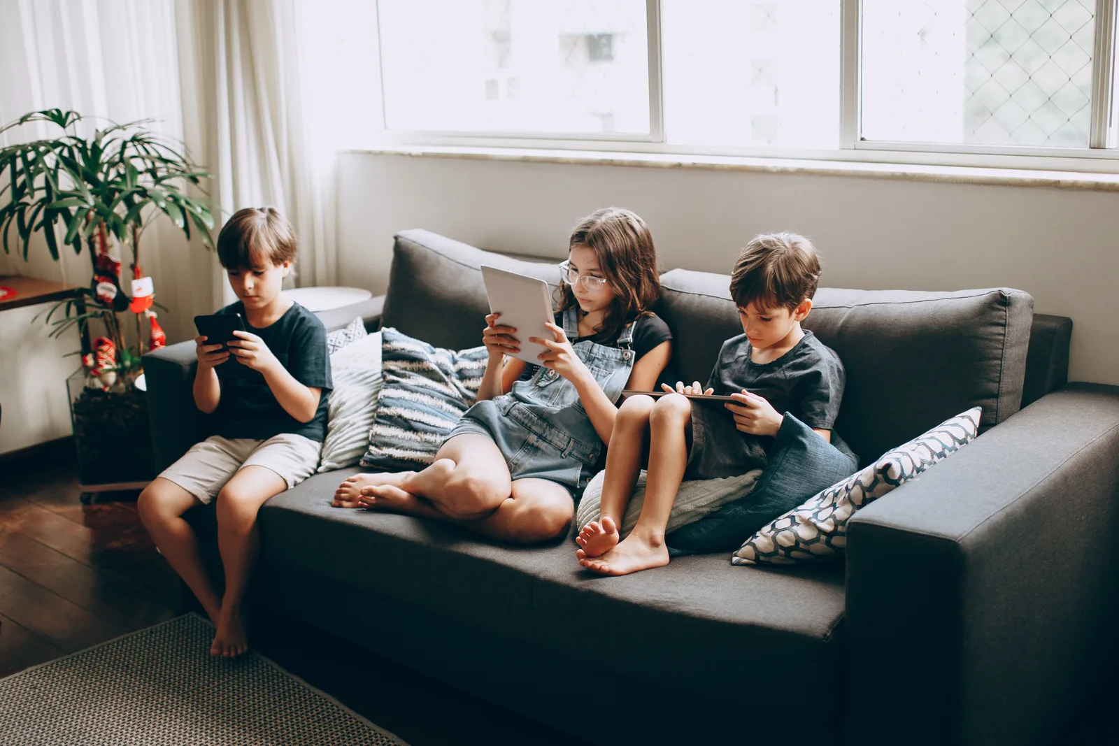 Three children on a gray sofa engrossed in digital devices, indoors. Family agreements changes: AI, tracking and in-app money