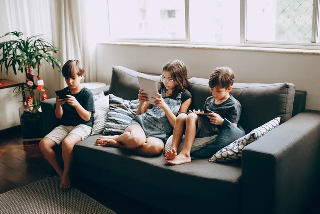 Three children on a gray sofa engrossed in digital devices, indoors. Family agreements changes: AI, tracking and in-app money