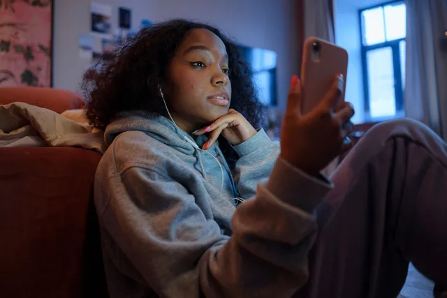 Teenage girl using smartphone indoors with earphones, wearing a hoodie, and looking relaxed. Teen social media use laws: finding the rule that applies